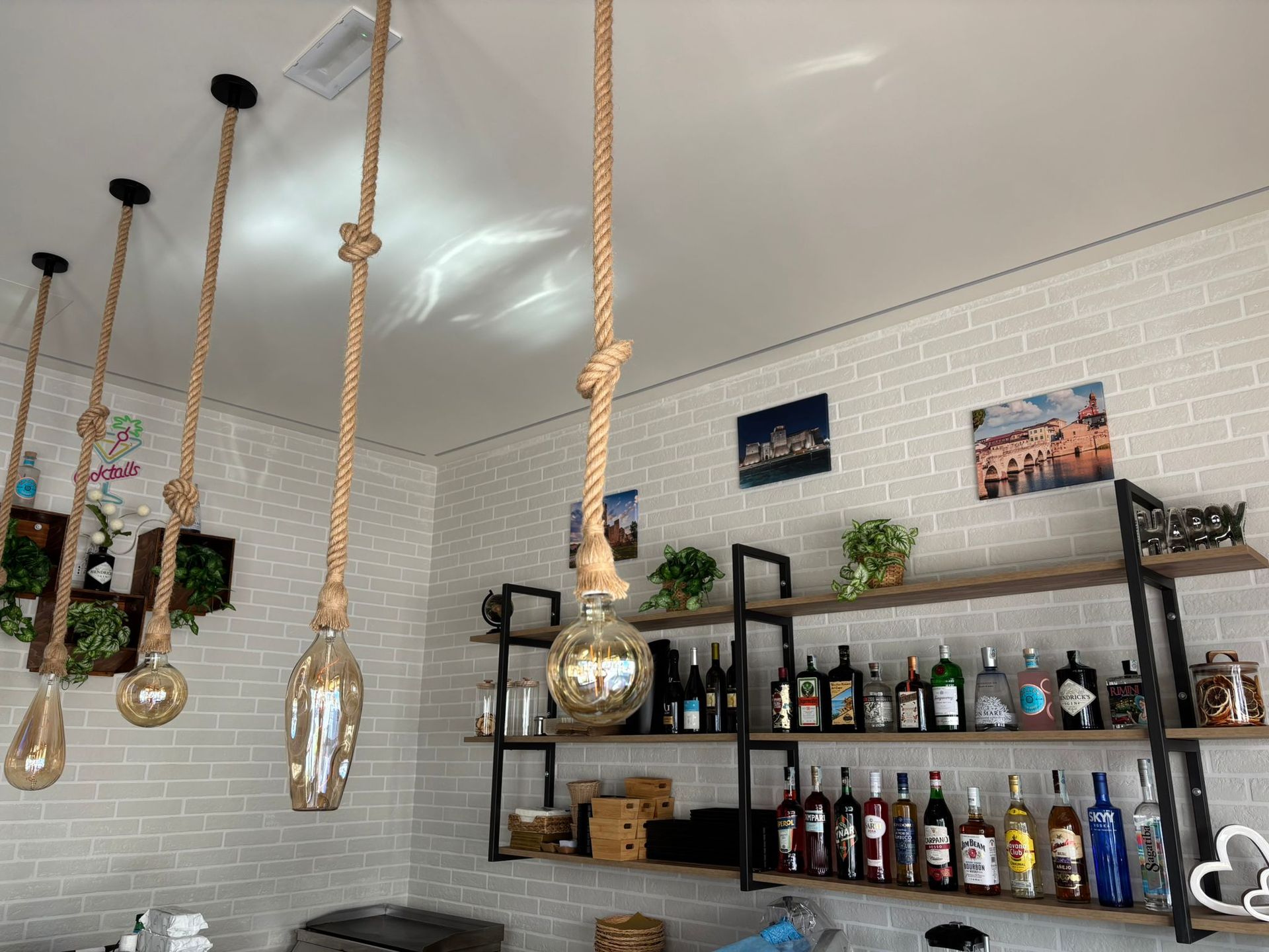 Un'area bar rustica con lampade a sospensione in corda, pareti in mattoni bianchi e scaffalature in legno a più livelli rifornite di liquori.