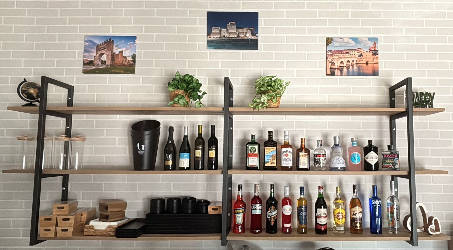 Mensole a muro fissate a una parete di mattoni, con file di bottiglie di liquore di vario tipo, bicchieri e piccole piante.