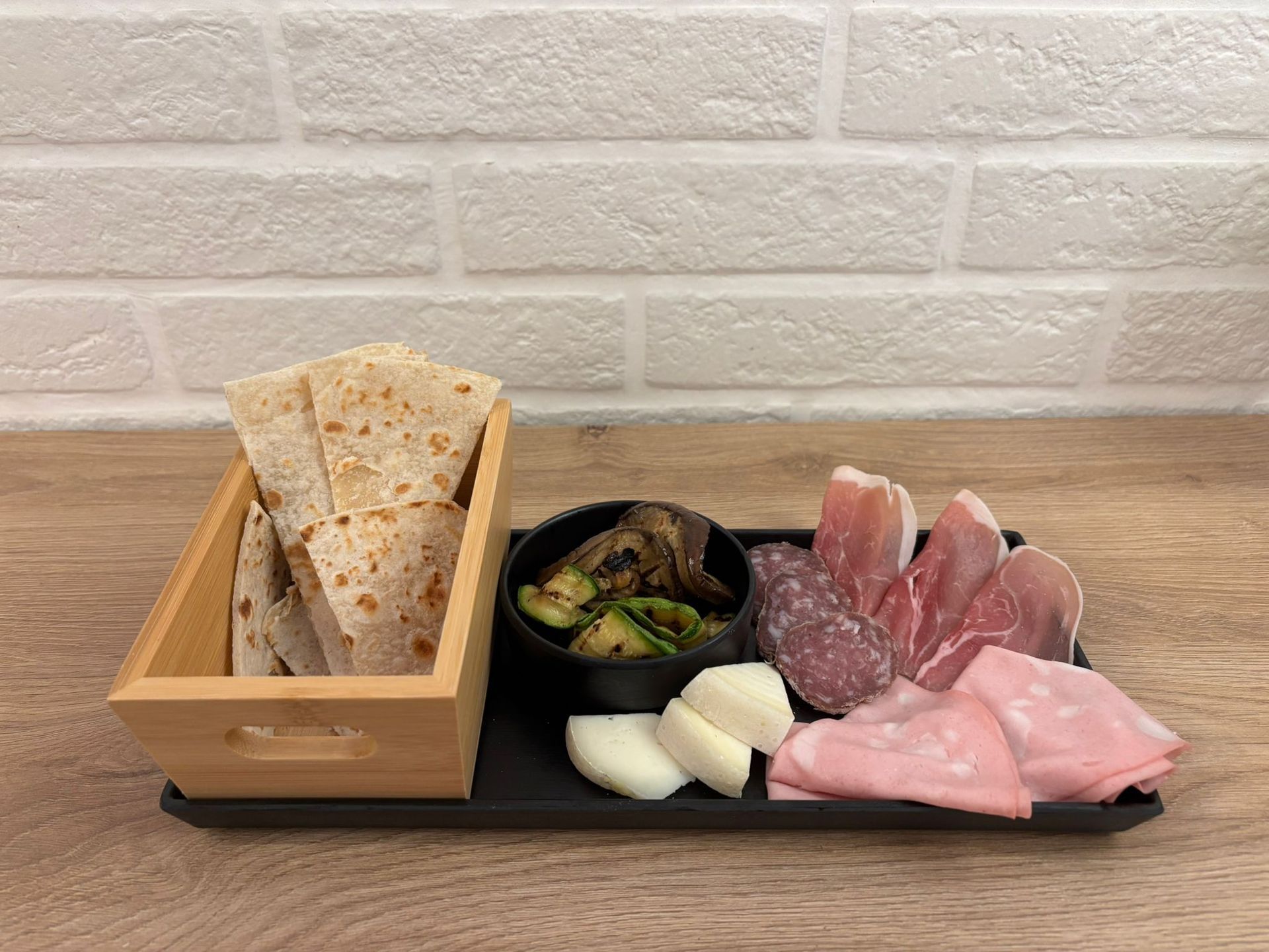 Un piatto da portata presenta una scatola di legno contenente pane piatto, verdure grigliate, salumi e formaggio su un vassoio nero appoggiato a una parete.
