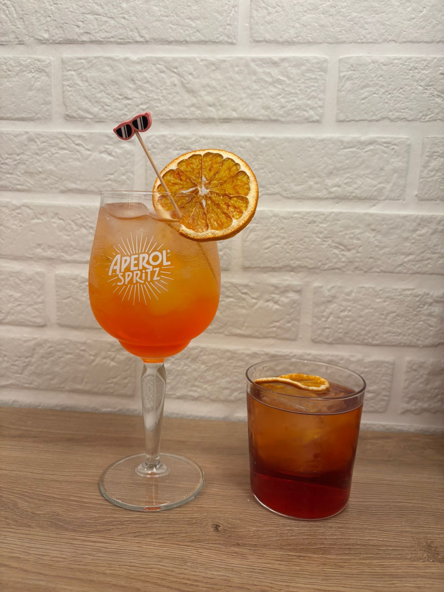 Un Aperol Spritz in un bicchiere di marca e un Negroni in un tumbler, entrambi guarniti con fette d'arancia essiccate su un tavolo di legno.