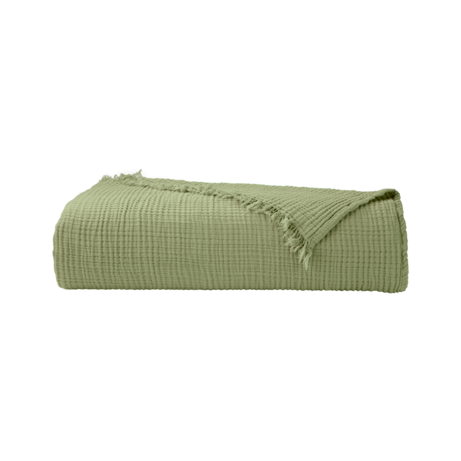 Un plaid en mousseline de coton texturée vert sauge, aux bords effilochés, soigneusement plié sur un fond blanc.