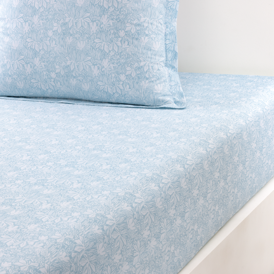 Drap-housse et taie d'oreiller bleu clair à motifs floraux botaniques blancs sur un cadre de lit devant un fond blanc.