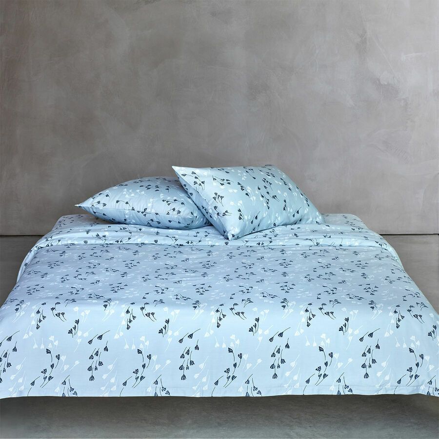 Un couvre-lit bleu clair et deux oreillers assortis ornés d'un délicat motif floral foncé, sur un mur gris.