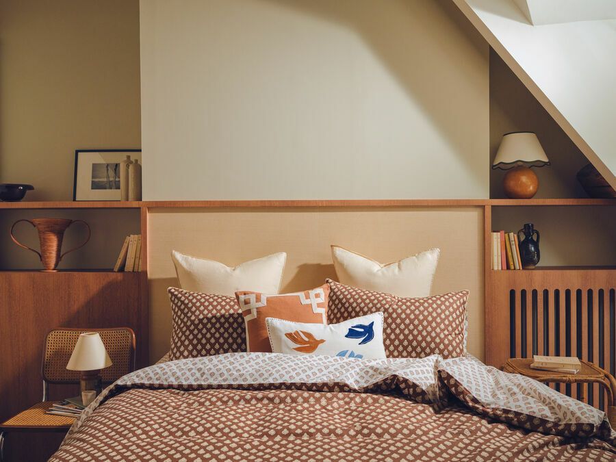 Une chambre avec un lit doté de draps marron à motifs, d'oreillers crème et d'étagères en bois intégrées sous un plafond mansardé.