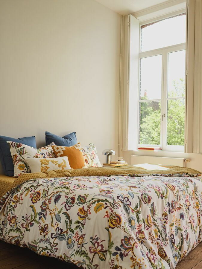Un lit avec une couette à motifs floraux colorés, des oreillers bleus et jaunes, à côté d'une fenêtre aux rideaux crème, dans une chambre lumineuse.