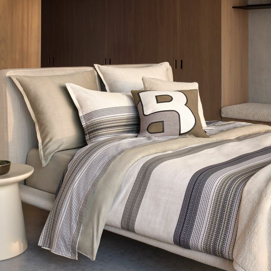 Un lit moderne avec une literie aux tons neutres, un jeté rayé et un coussin décoratif orné d'une grande lettre B.