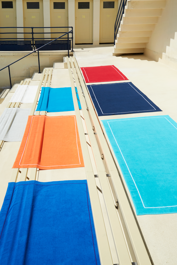 Des serviettes rectangulaires colorées sont soigneusement disposées sur un escalier en béton devant des cabines de plage aux tons clairs.
