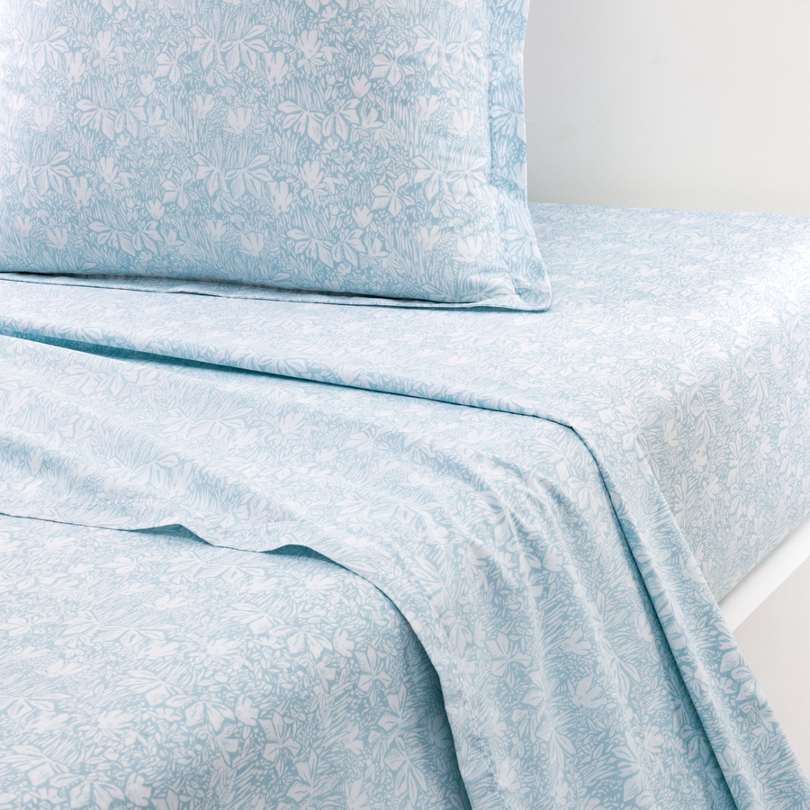 Un oreiller et des draps pliés à motifs floraux bleu clair et blancs se détachent sur un fond blanc uni.