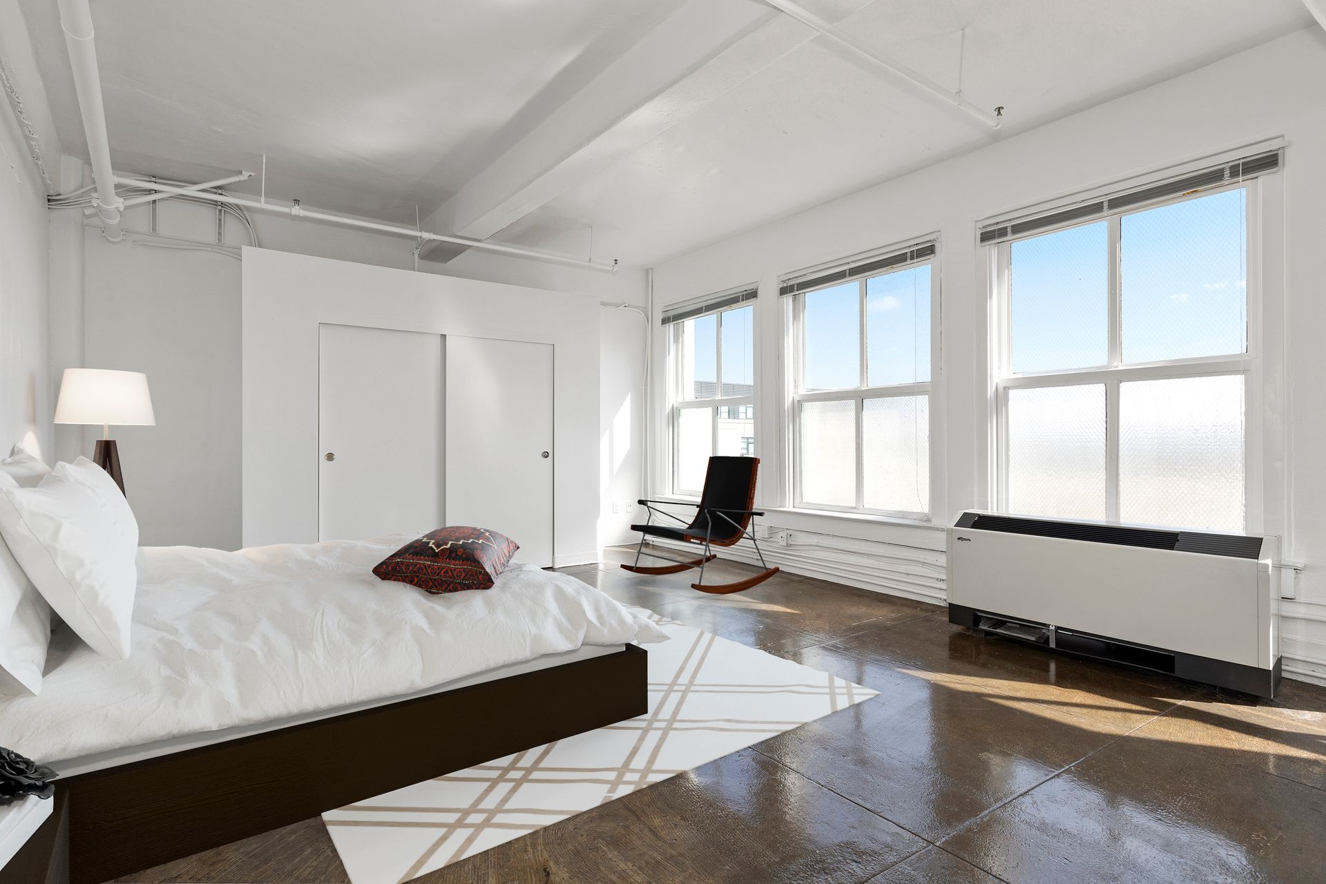 Bright white bedroom with large windows, a bed, chair, and dark hardwood floor
