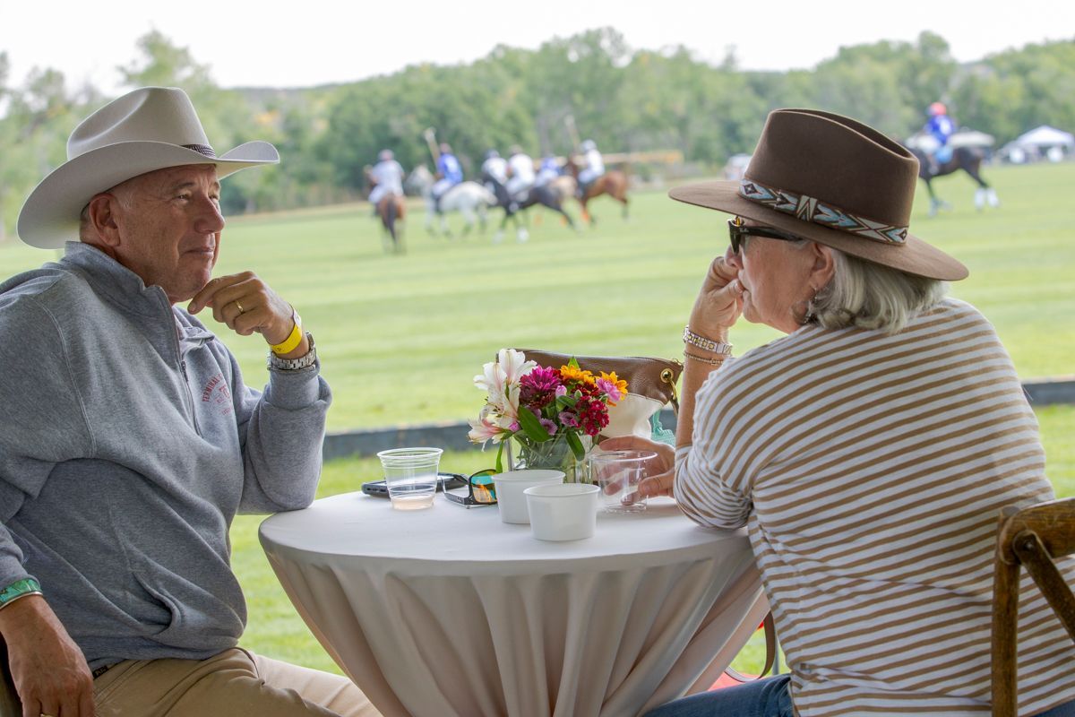 Two people in hats seated at a table, watching a polo match.