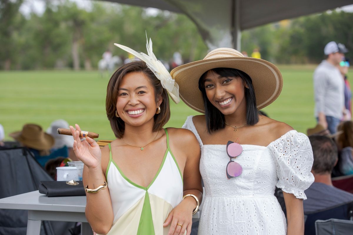 Two women smile, one with a cigar, at an outdoor event. One wears a sun hat and white dress.