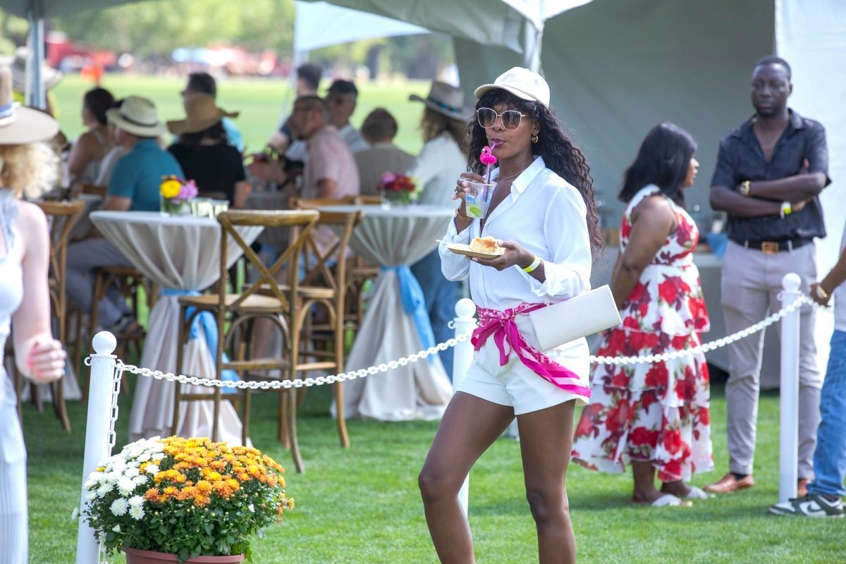 Woman in white shorts and top sips a drink at an outdoor event with others.