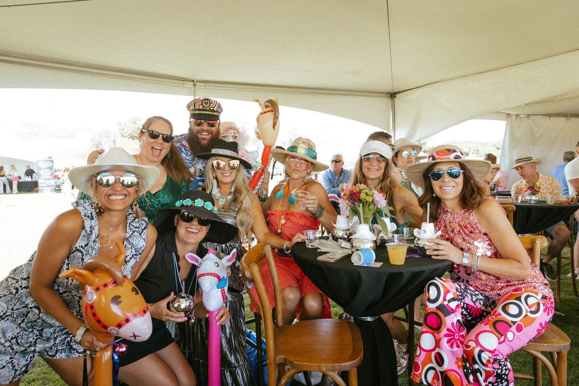 Group of people in hats and colorful outfits gather outdoors at a party under a tent.