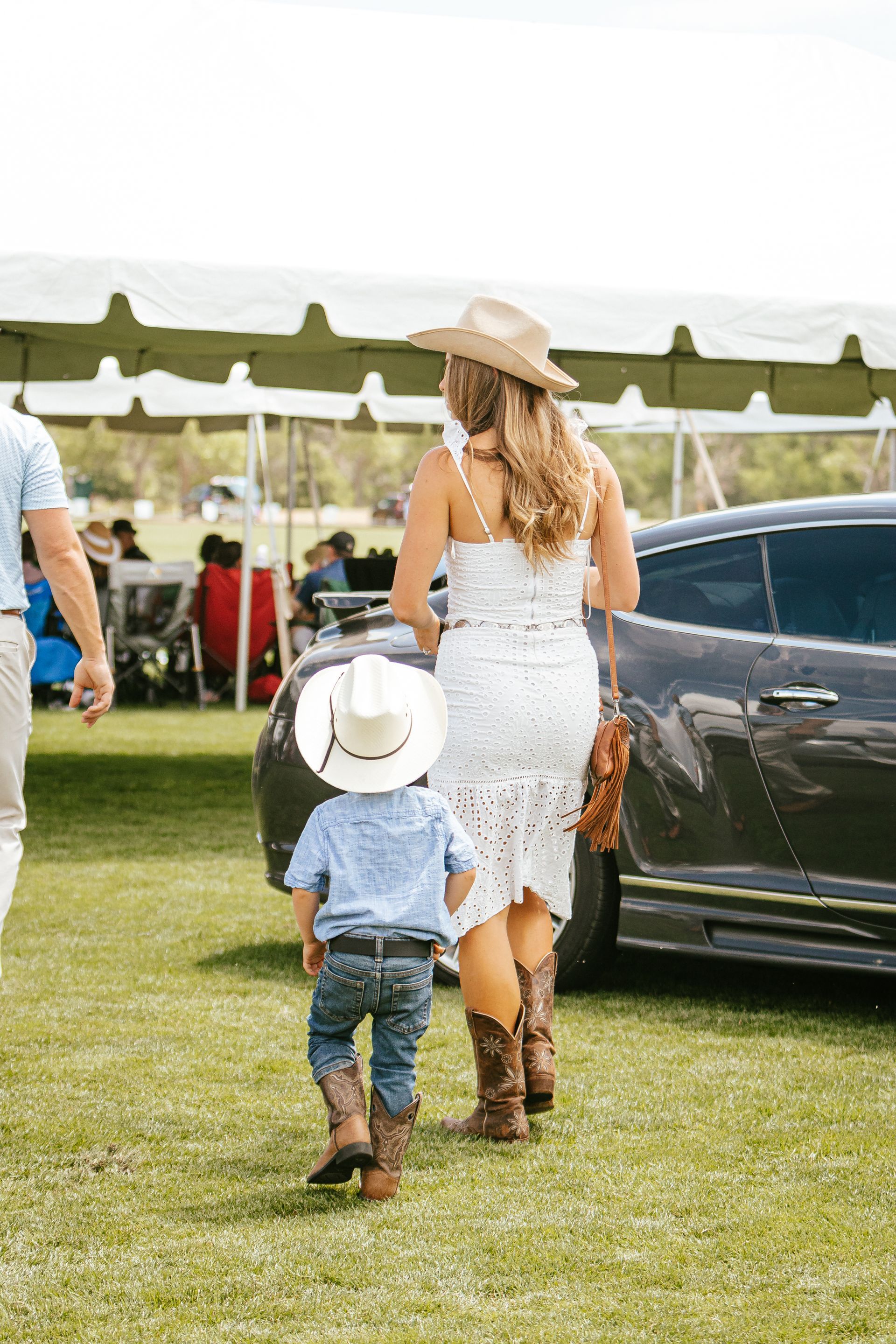 Woman and young child, both in cowboy hats and boots, walking on grass near a car and event tent.