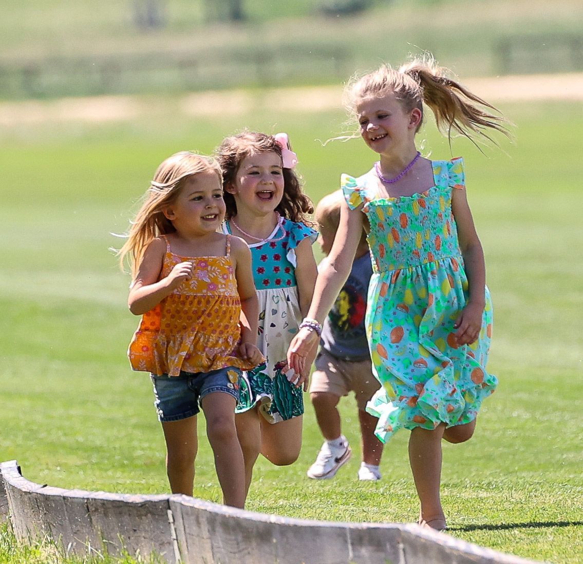 Children running on grass near a fence, laughing. Two girls in dresses, one in shorts.