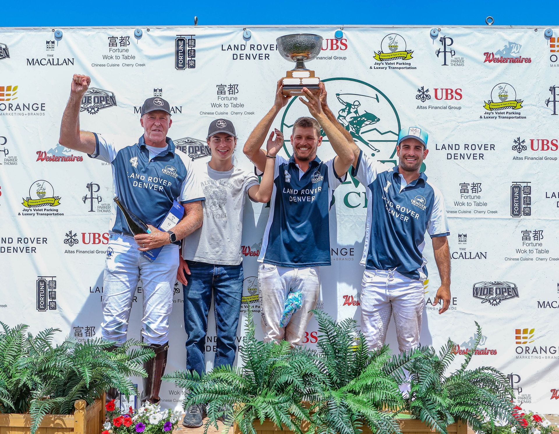 Polo team celebrating victory, holding trophy. Men in team jerseys, smiling, standing on a podium outdoors.