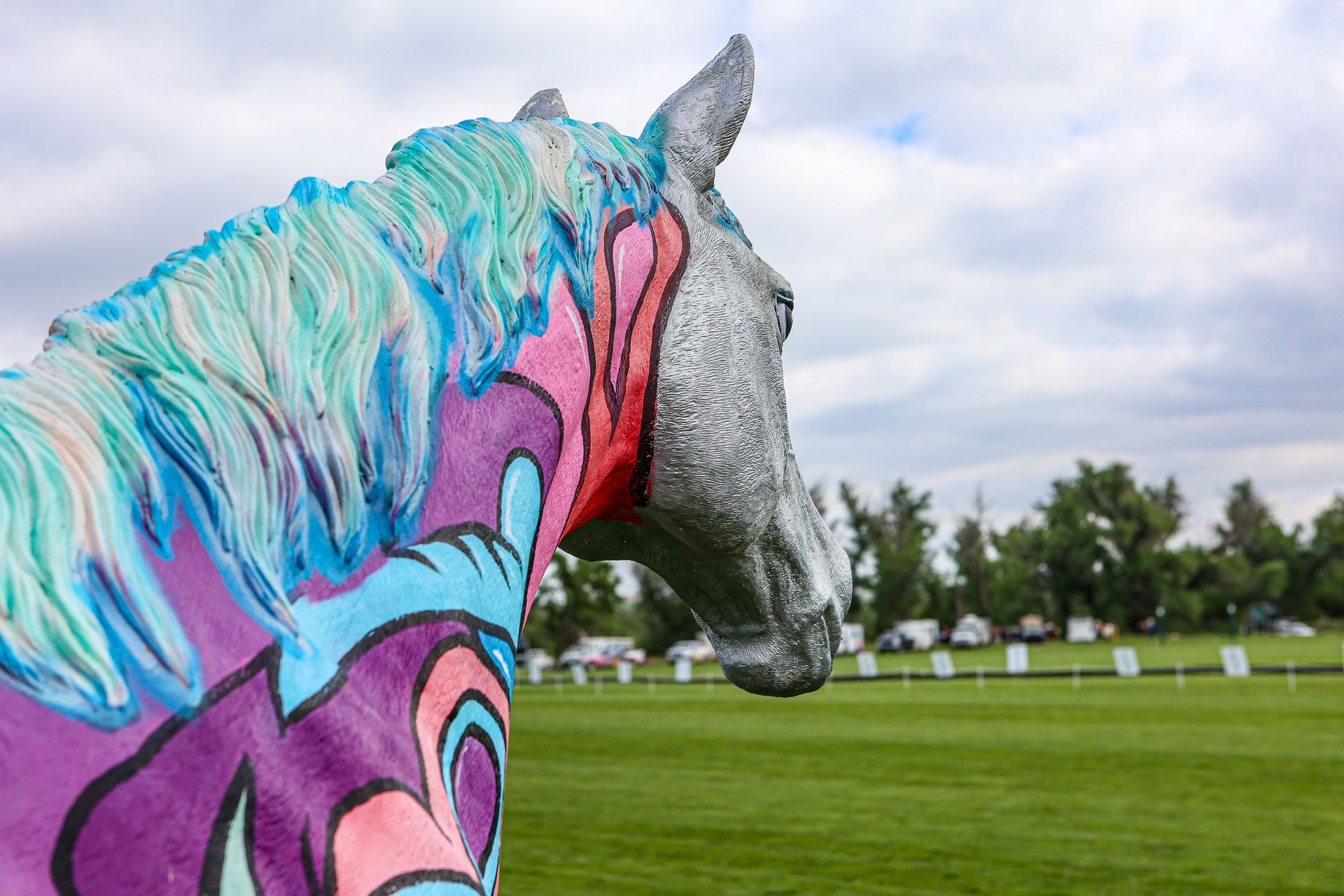 Painted horse statue on green field, colorful mane and body art, cloudy sky background.
