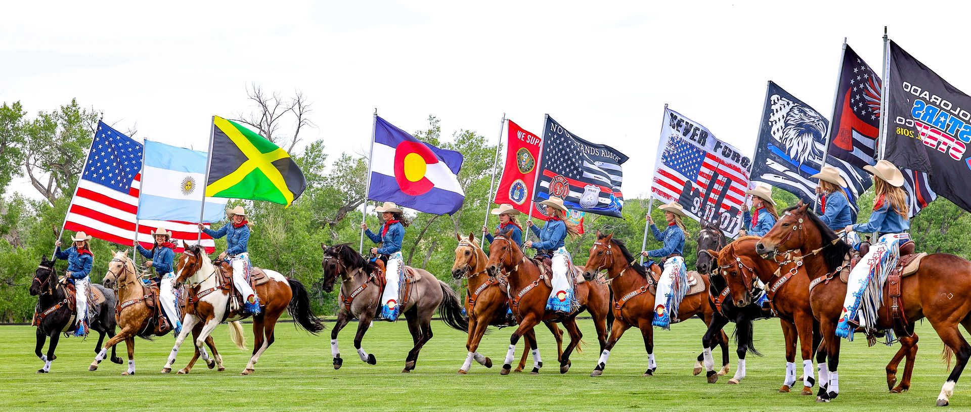 People on horseback holding flags, including the US, Colorado, and Jamaican flags, on a grassy field.