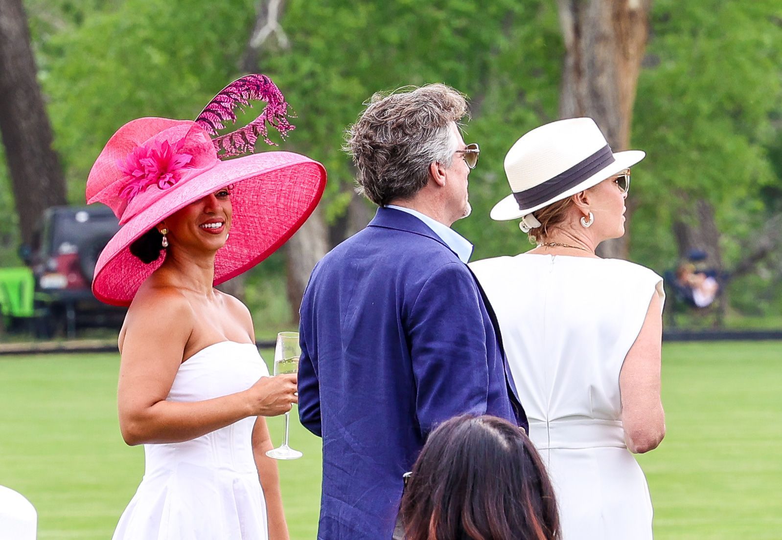 Woman in a pink hat, two people in hats and a white dress, standing on grass.