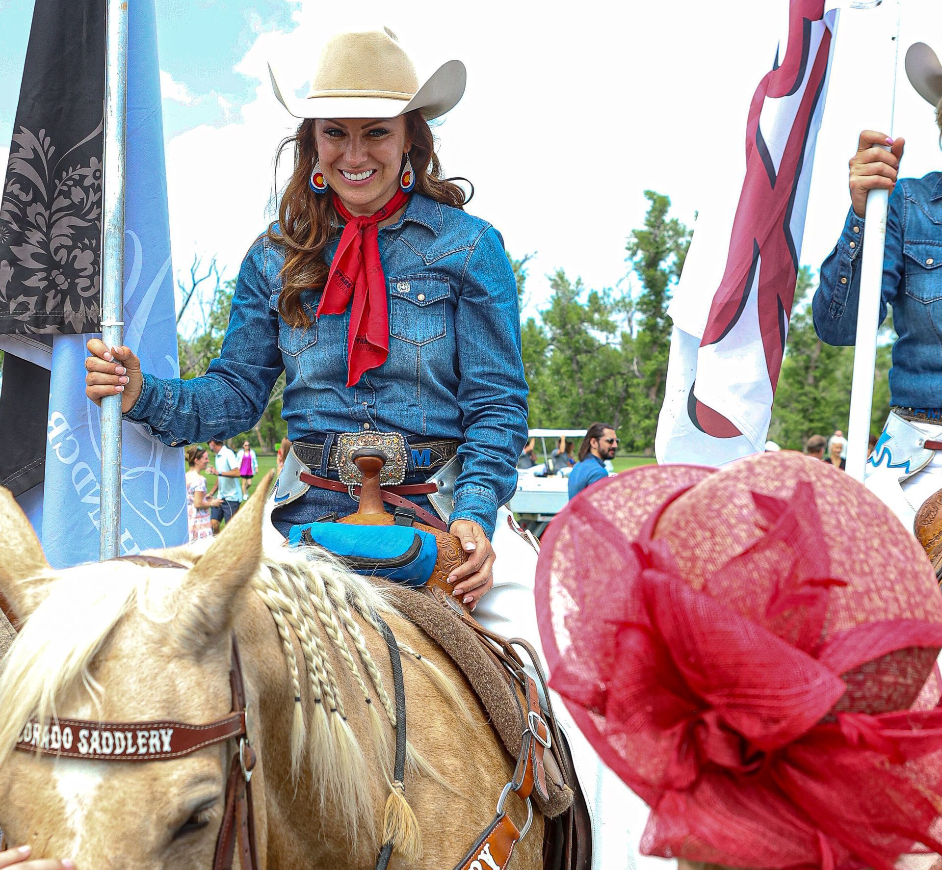 Cowgirl on horseback smiles, holding a blue flag. She wears denim, a red scarf, and a tan cowboy hat.