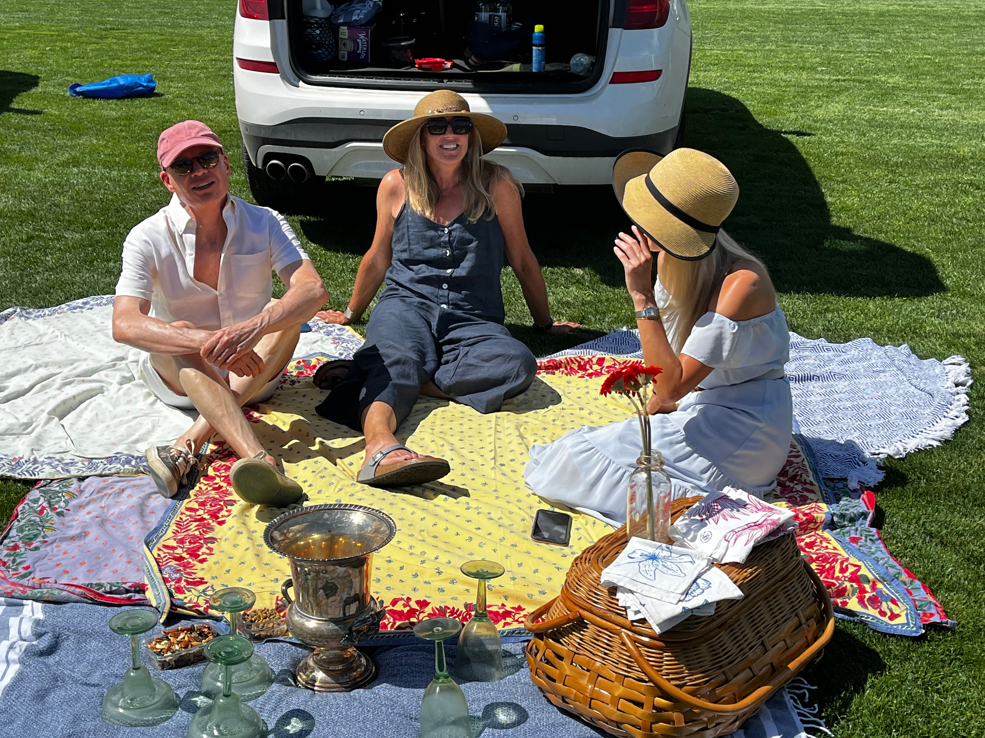 A group of people are sitting on a blanket in front of a car.