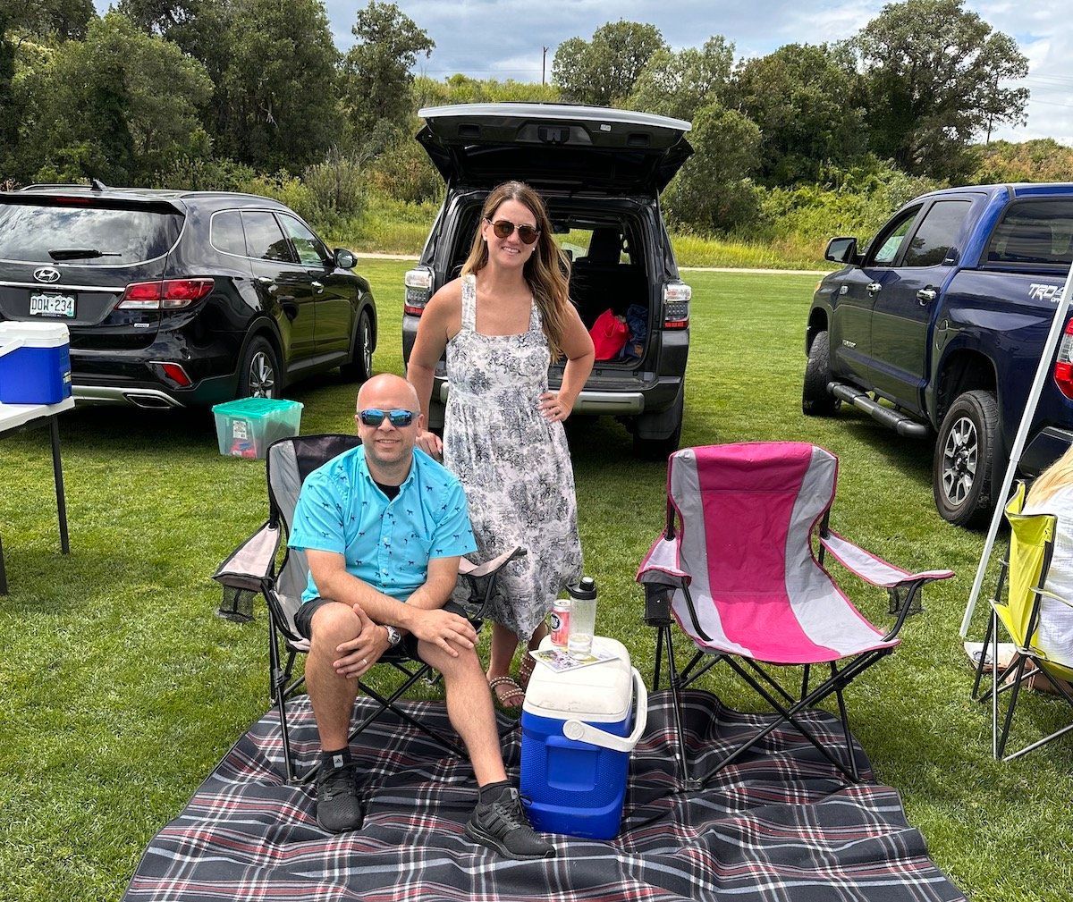 A man and a woman are sitting on a blanket in front of a car.