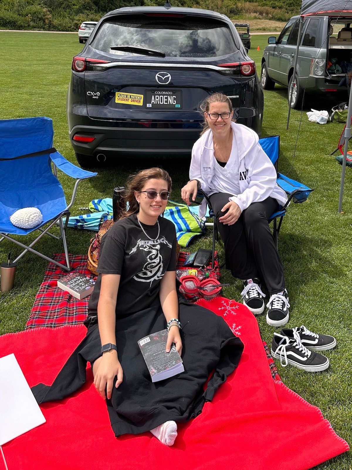 Two women are sitting on a red blanket in front of a car.