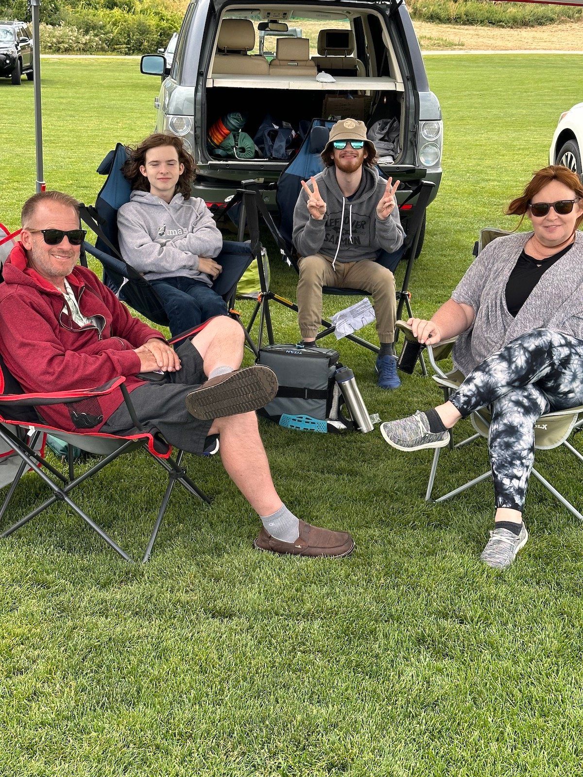 A group of people are sitting in chairs in the grass in front of a car.