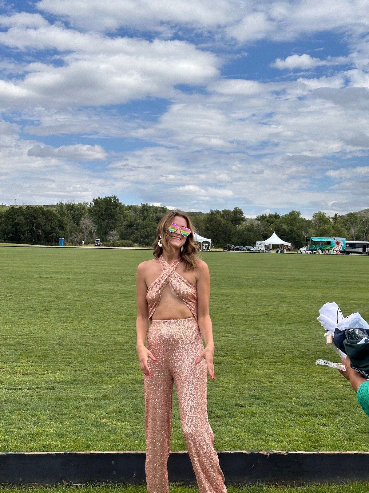 A woman in a sequined jumpsuit is standing in a grassy field.