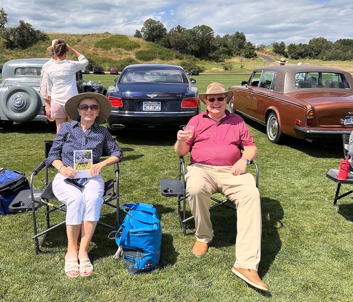 A man and a woman are sitting in chairs in a field with cars in the background.