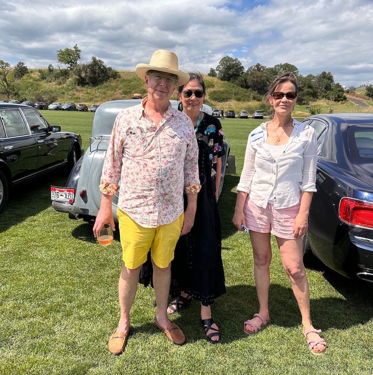 Three people are posing for a picture in front of a car in a field.