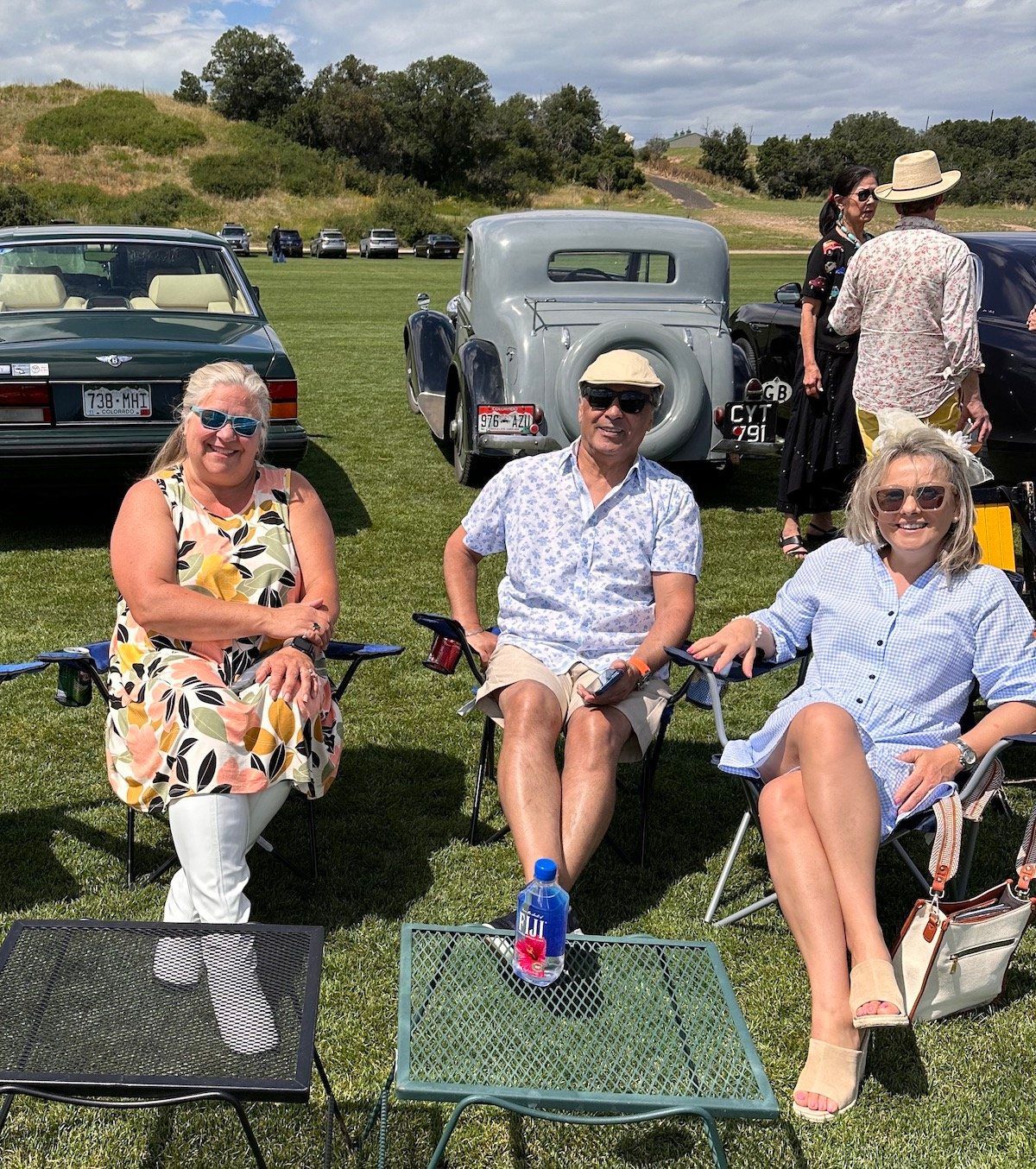Three people are sitting in chairs in a field with cars in the background.