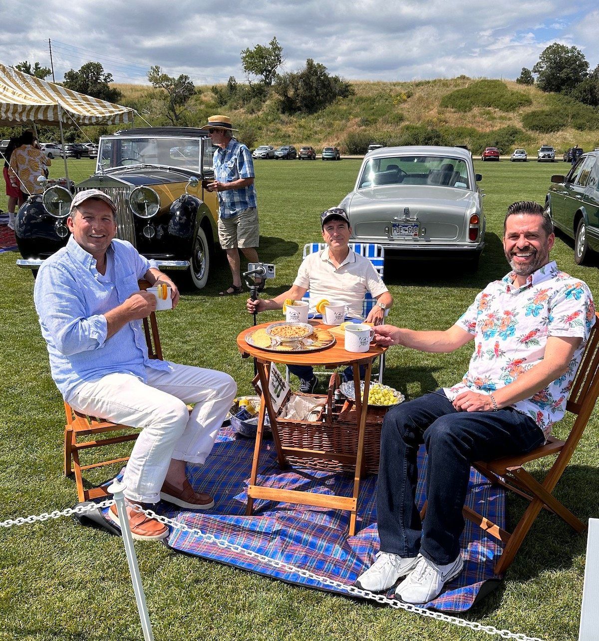 A group of men are having a picnic in a field with cars in the background.