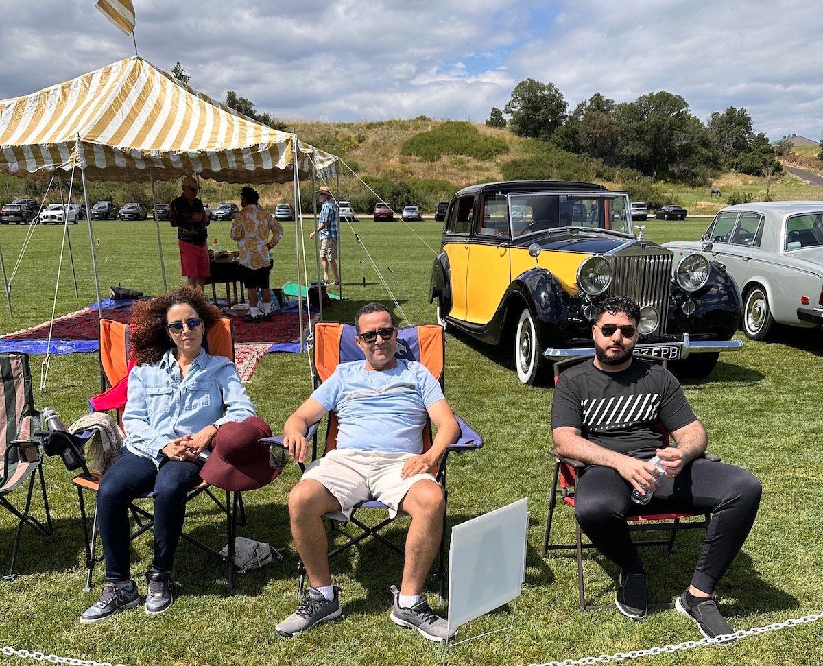 A group of people are sitting in chairs in a field in front of old cars.