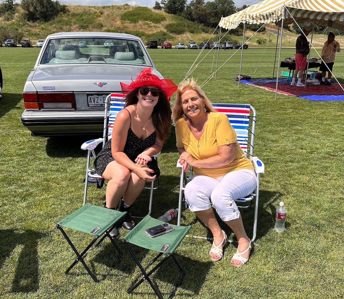 Two women are sitting in chairs in a field next to a car.