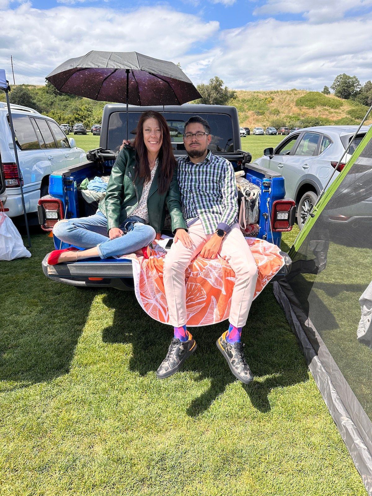 A man and a woman are sitting in the back of a truck under an umbrella.