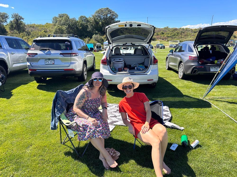 Two women are sitting in chairs in a field with cars parked in the background.