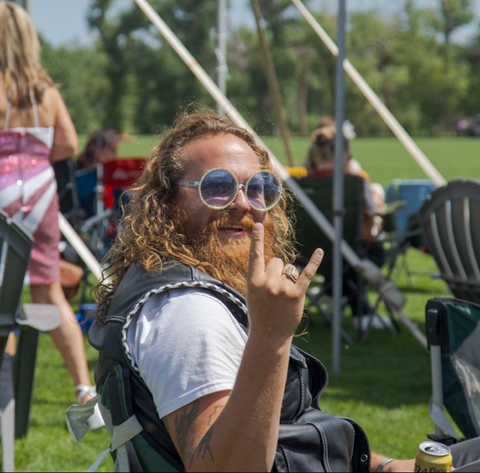 Man with long hair and beard giving a rock on hand gesture, wearing sunglasses and a leather vest, outside.