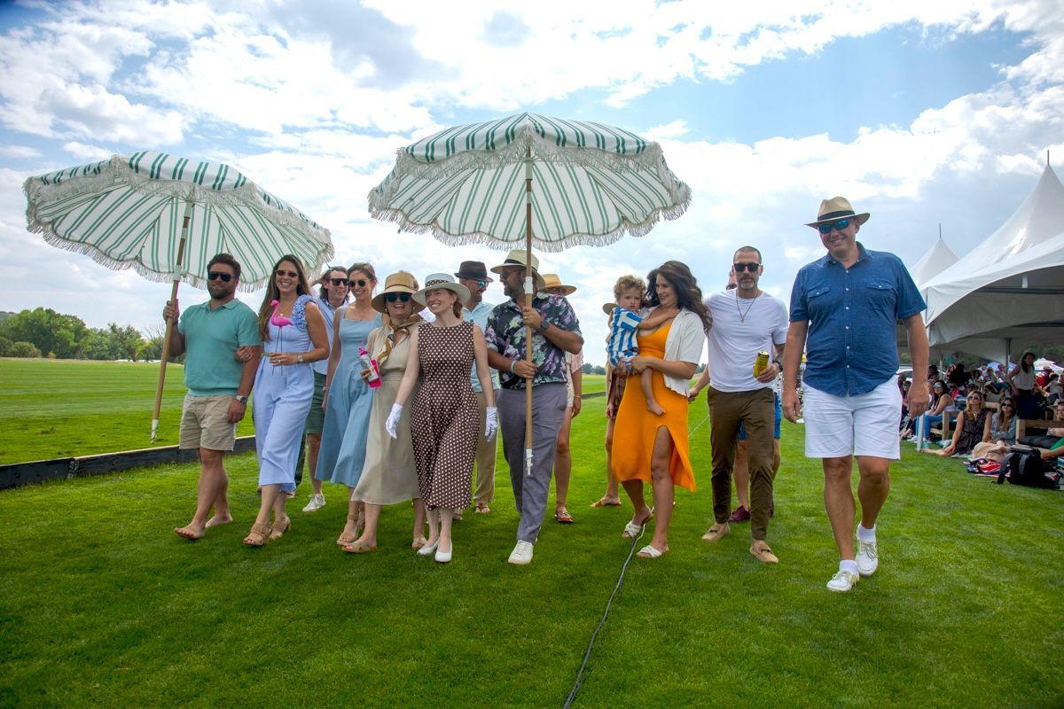 People walking on grass, shaded by parasols at an outdoor event. Bright sky, tent in background.