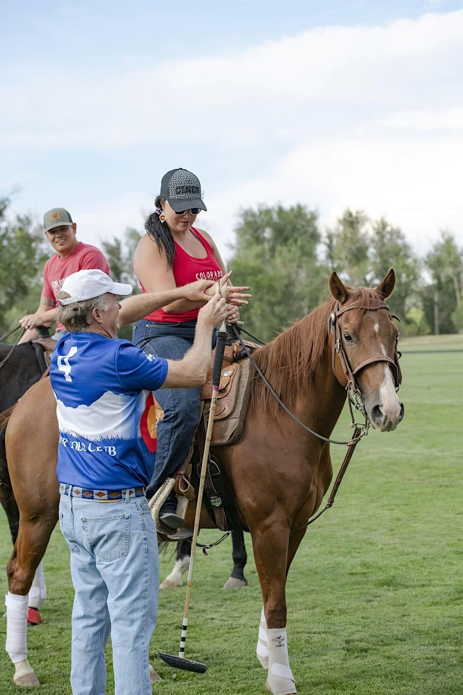 Man in blue shirt assisting woman on brown horse, polo mallet in hand, outdoors.