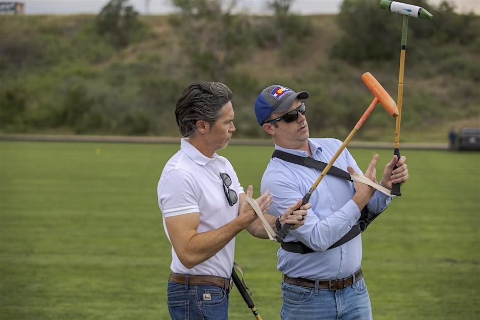 Two men on a polo field, one holding a mallet, the other demonstrating its use, both wear casual clothing.