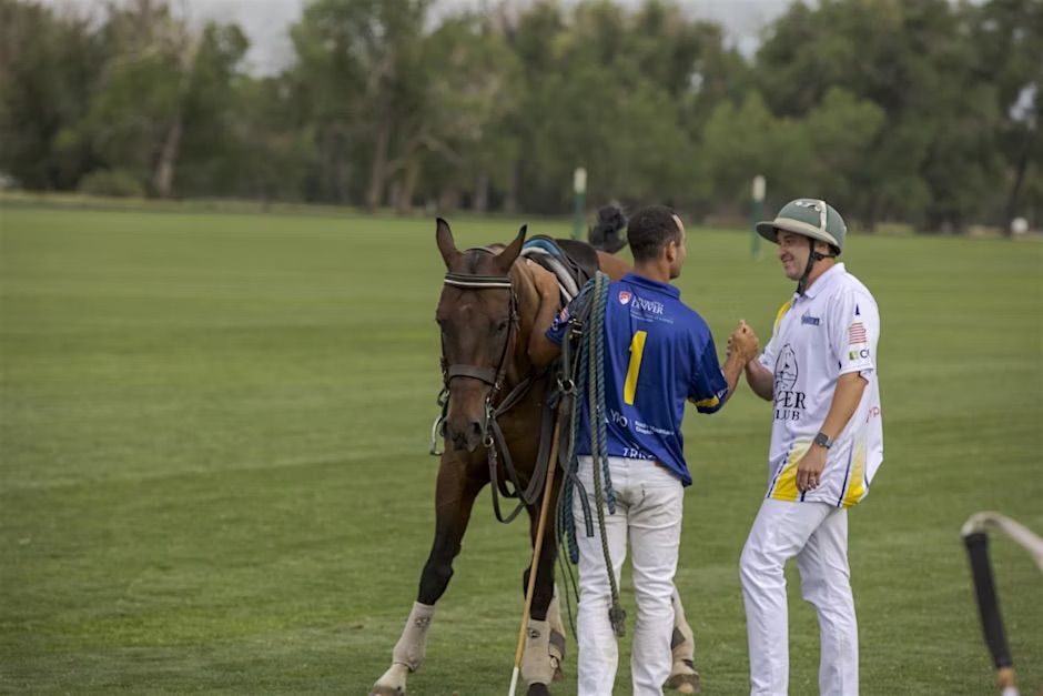 Two polo players fist bump next to a horse on a grassy field.