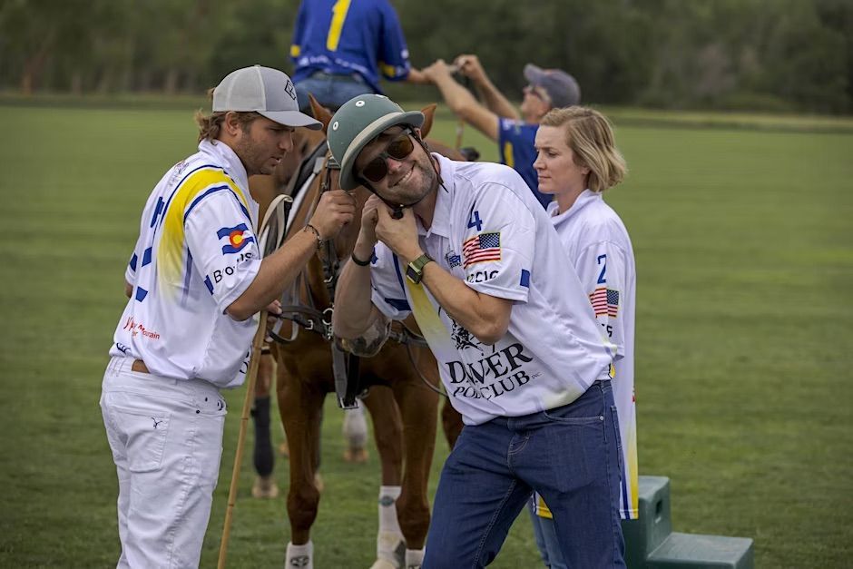Polo players in white shirts, one with a helmet, stand near a horse, with a helper in the background.