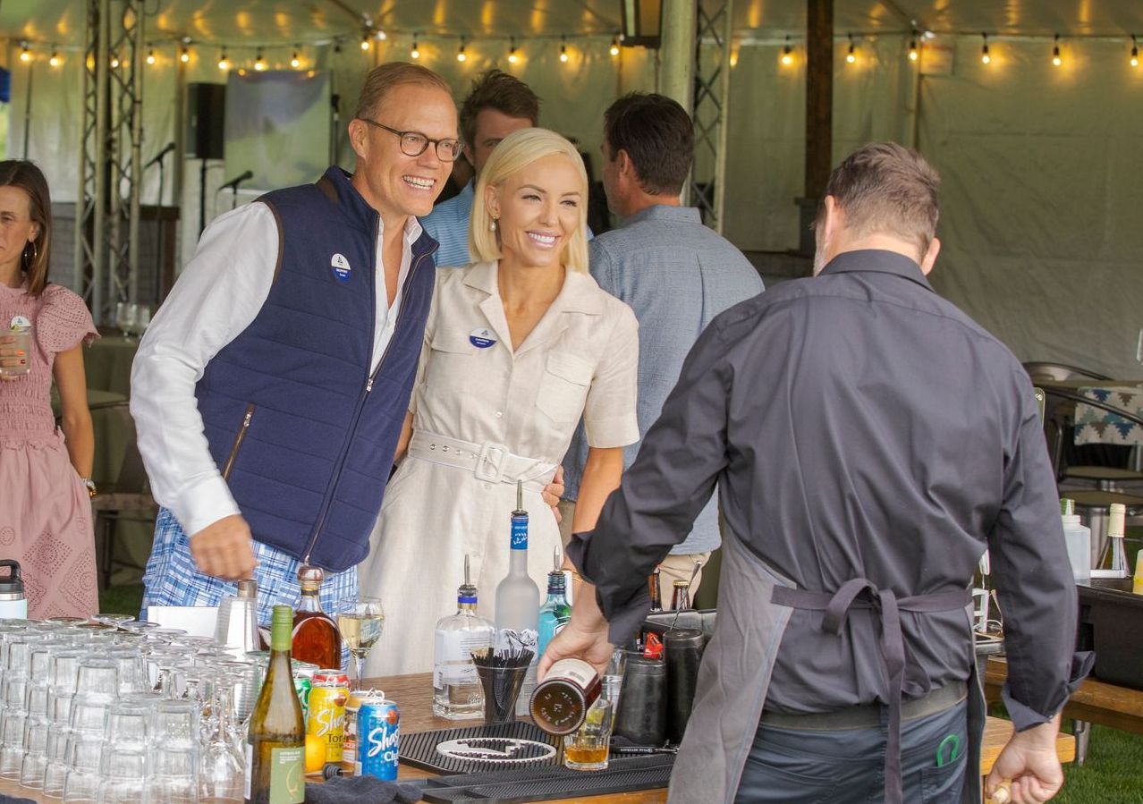 Man and woman at a bar smiling, bartender pouring a drink outdoors, other people in the background.