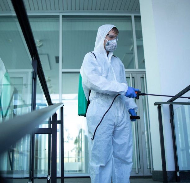 Una persona con traje protector blanco y mascarilla rocía desinfectante en un pasillo interior moderno.