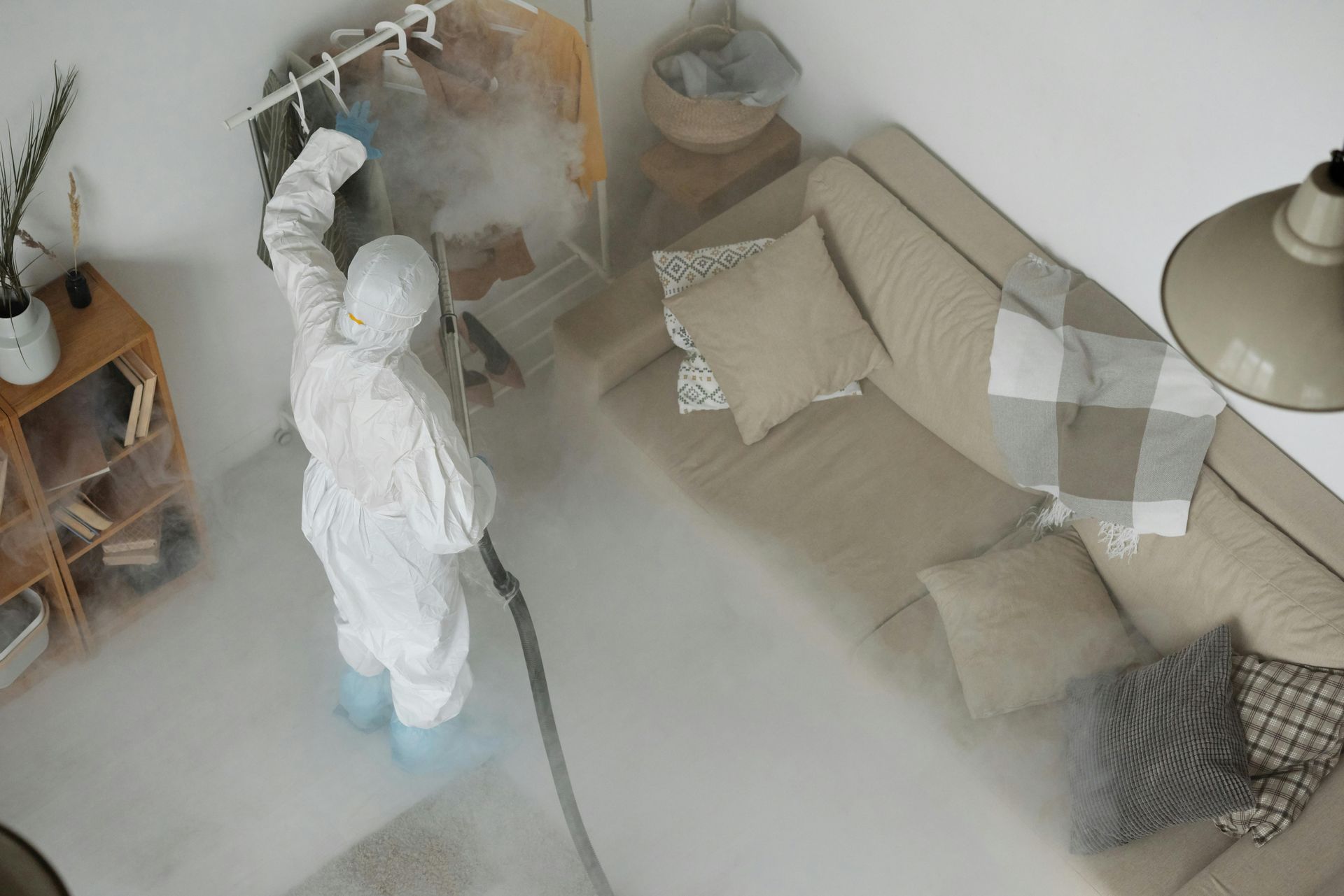 Una persona con traje protector desinfecta una sala de estar con una pistola pulverizadora, produciendo una niebla visible.