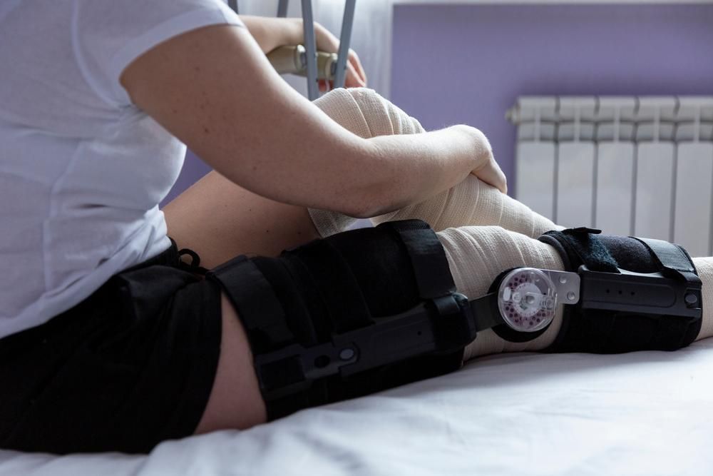 A Woman with A Knee Brace Is Sitting on A Bed — St Vincents Physiotherapy & Day Rehab in East Lismore, NSW