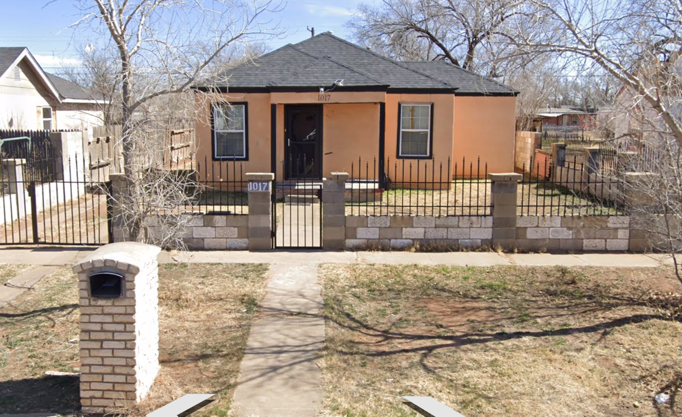 Small tan house with a front porch, black fence, and trees in a residential yard.