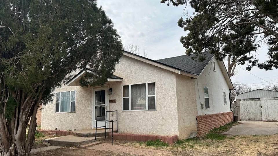 Small beige house with a front stoop, trees in front, and a white garage or shed beside it.