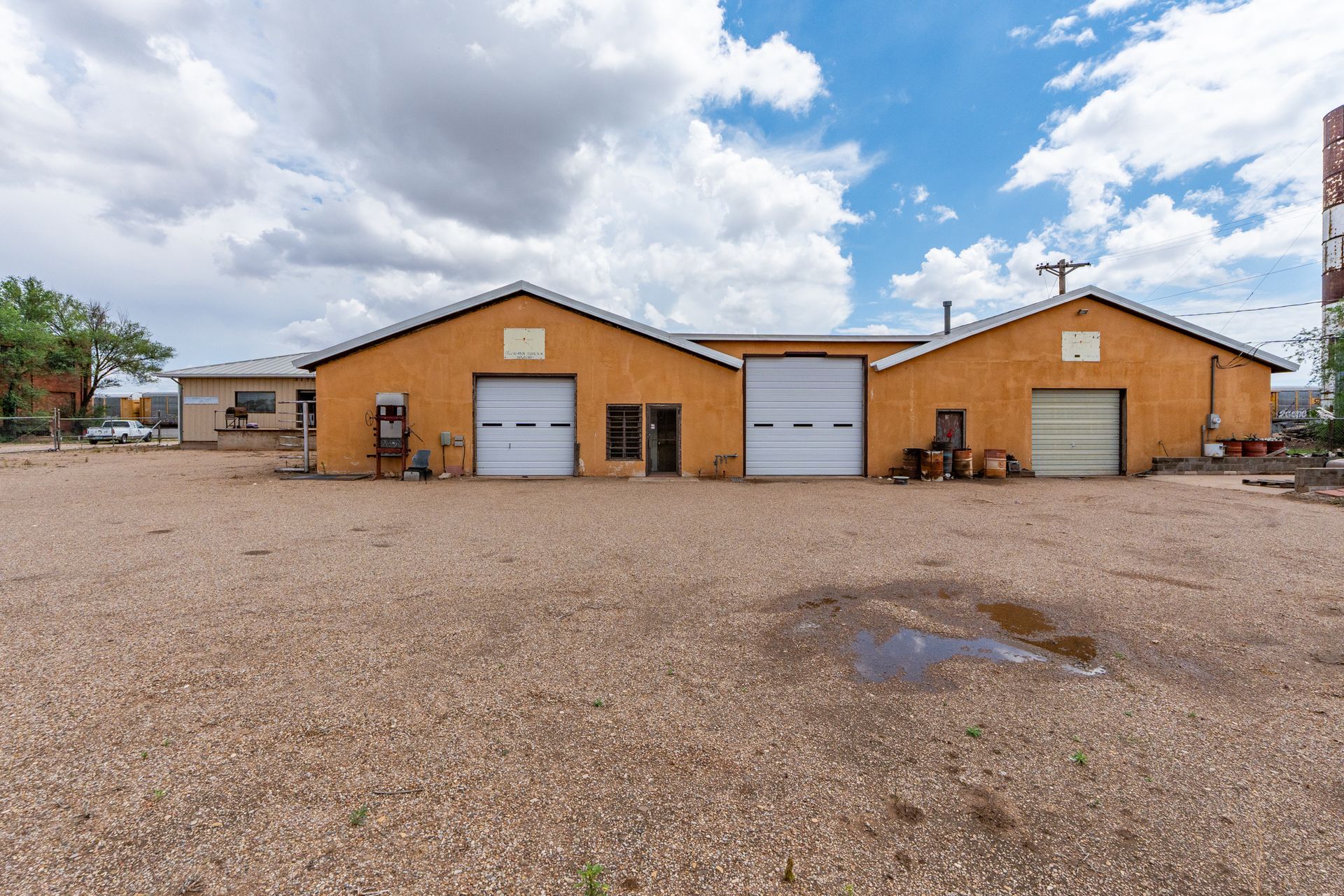 Two tan metal buildings with garage doors on a gravel lot under a cloudy sky